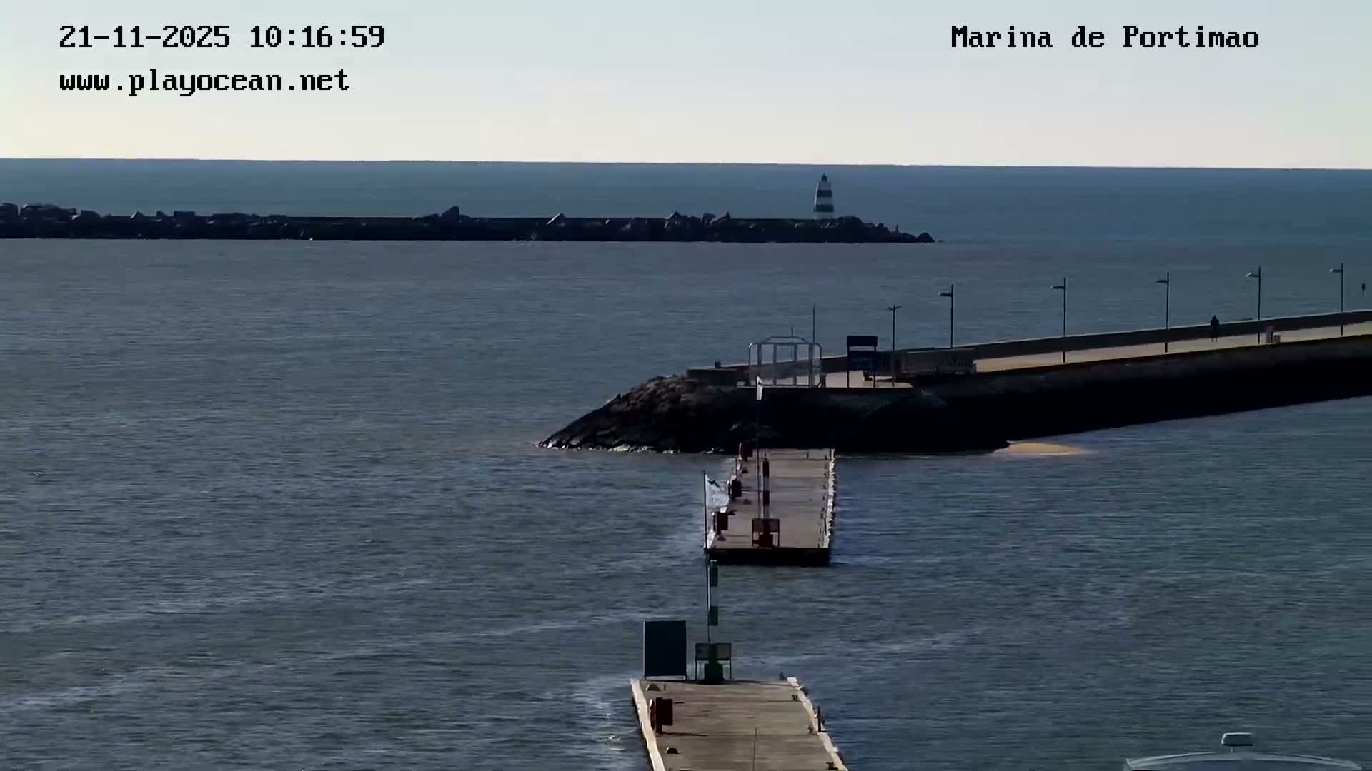 A clear and sunny sky overlooks calm blue-grey waters featuring multiple piers and jetties, including a large concrete pier with streetlights, and a distant striped lighthouse at the end of a long rocky breakwater.