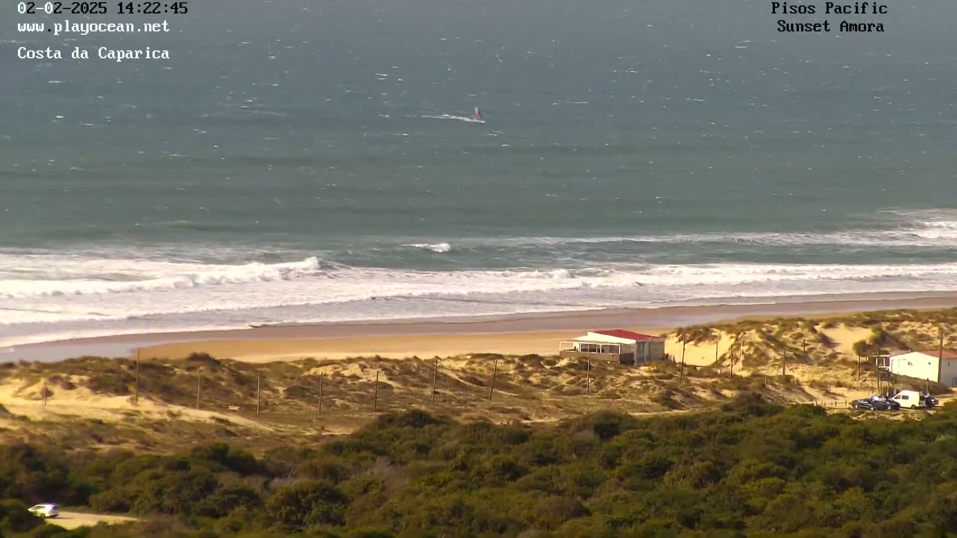 An ocean beach with moderate waves, a small building on the sand dunes, and a windsurfer in the distance under a partly sunny sky.