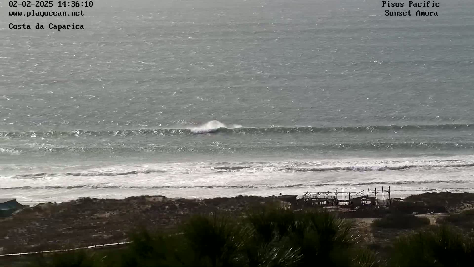 An ocean beach with waves breaking, seen from a high vantage point overlooking a sandy shore and some structures.