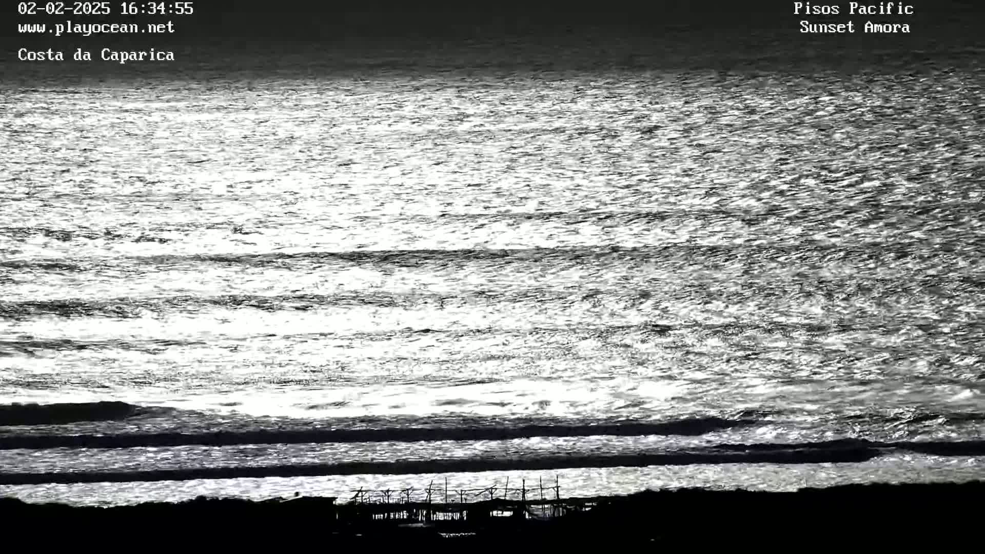 A mostly calm ocean with small waves reflecting sunlight, and a dark silhouette of a structure on the shore.