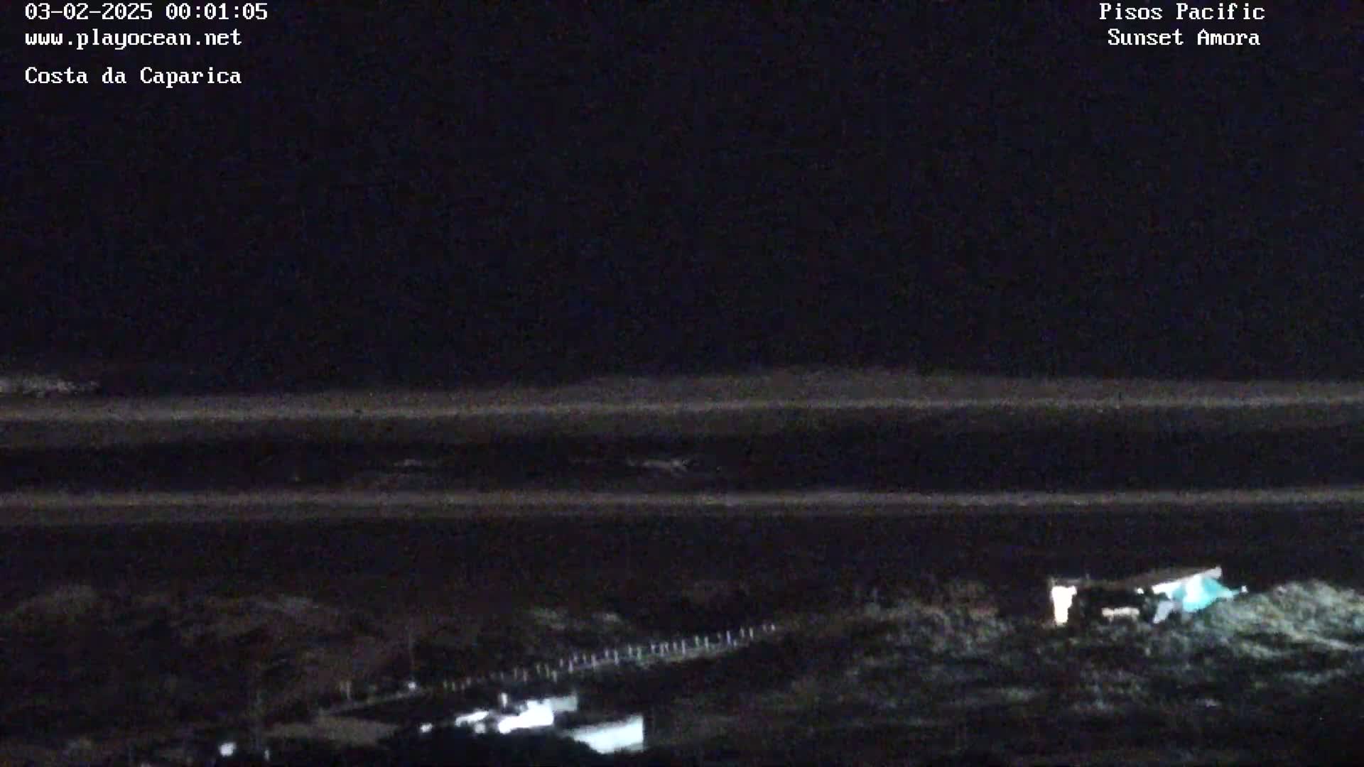 A dark, nighttime view of a beach with a calm ocean and a few lights visible on the shore.