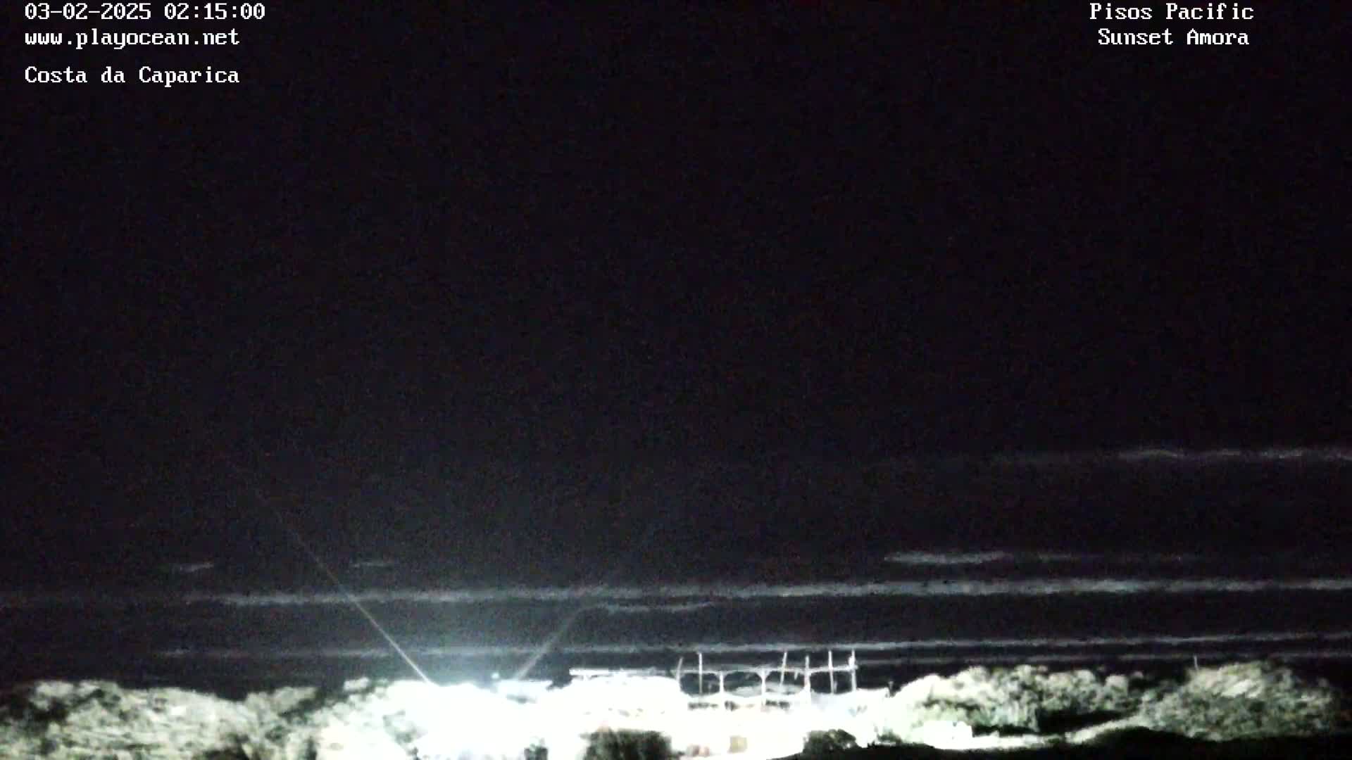 A nighttime coastal scene shows dark water under a mostly clear, dark sky with a few thin clouds, and faintly lit structures near the water's edge.