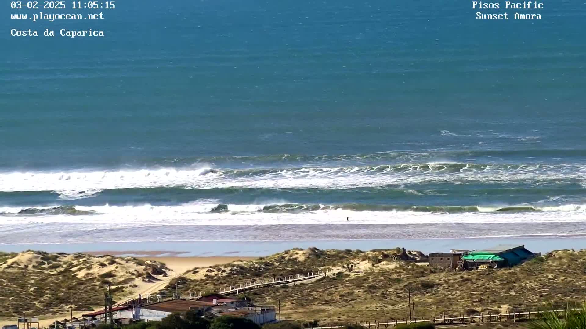 An elevated view shows a sandy beach with moderate waves breaking in the ocean under a clear sky.