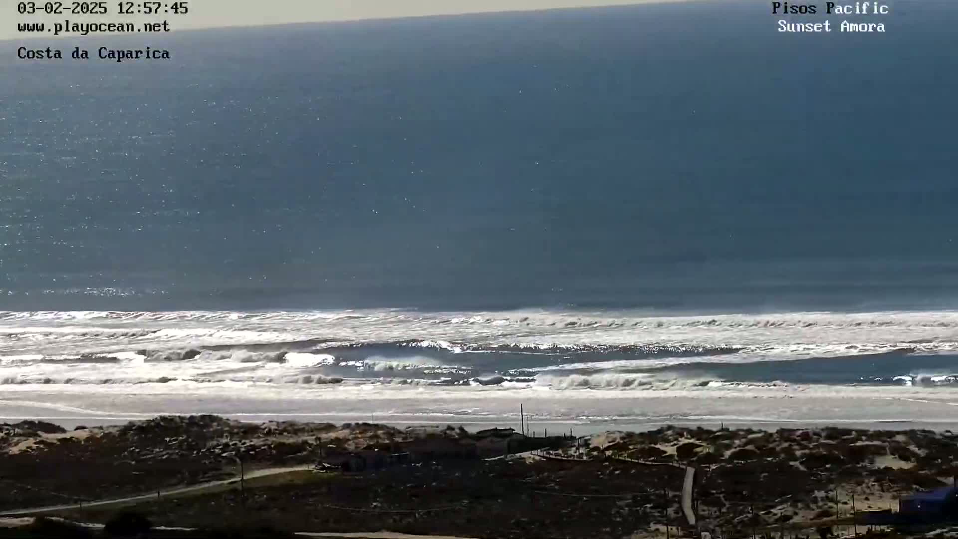 A high-angle view shows a sandy beach with moderate waves breaking on the shore under a mostly clear sky.