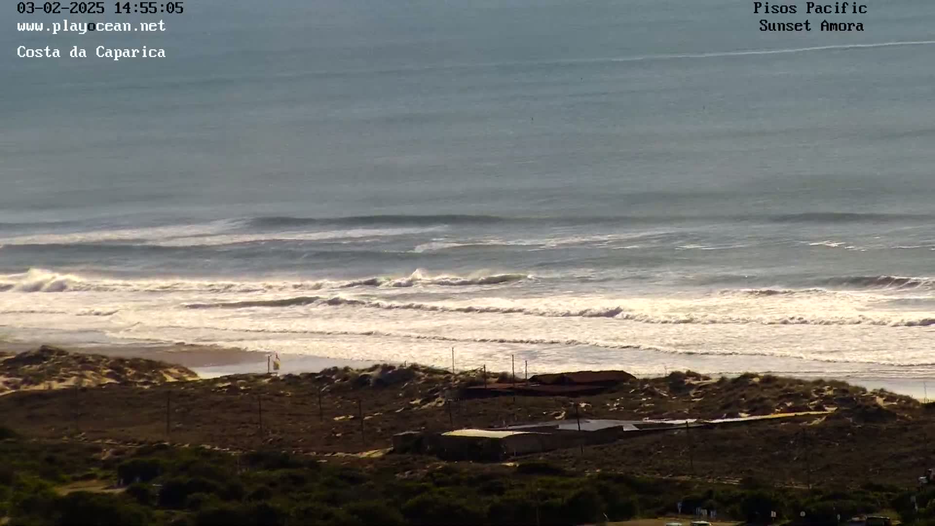Ocean waves roll onto a sandy beach near a low-lying building complex under a bright, sunny sky.