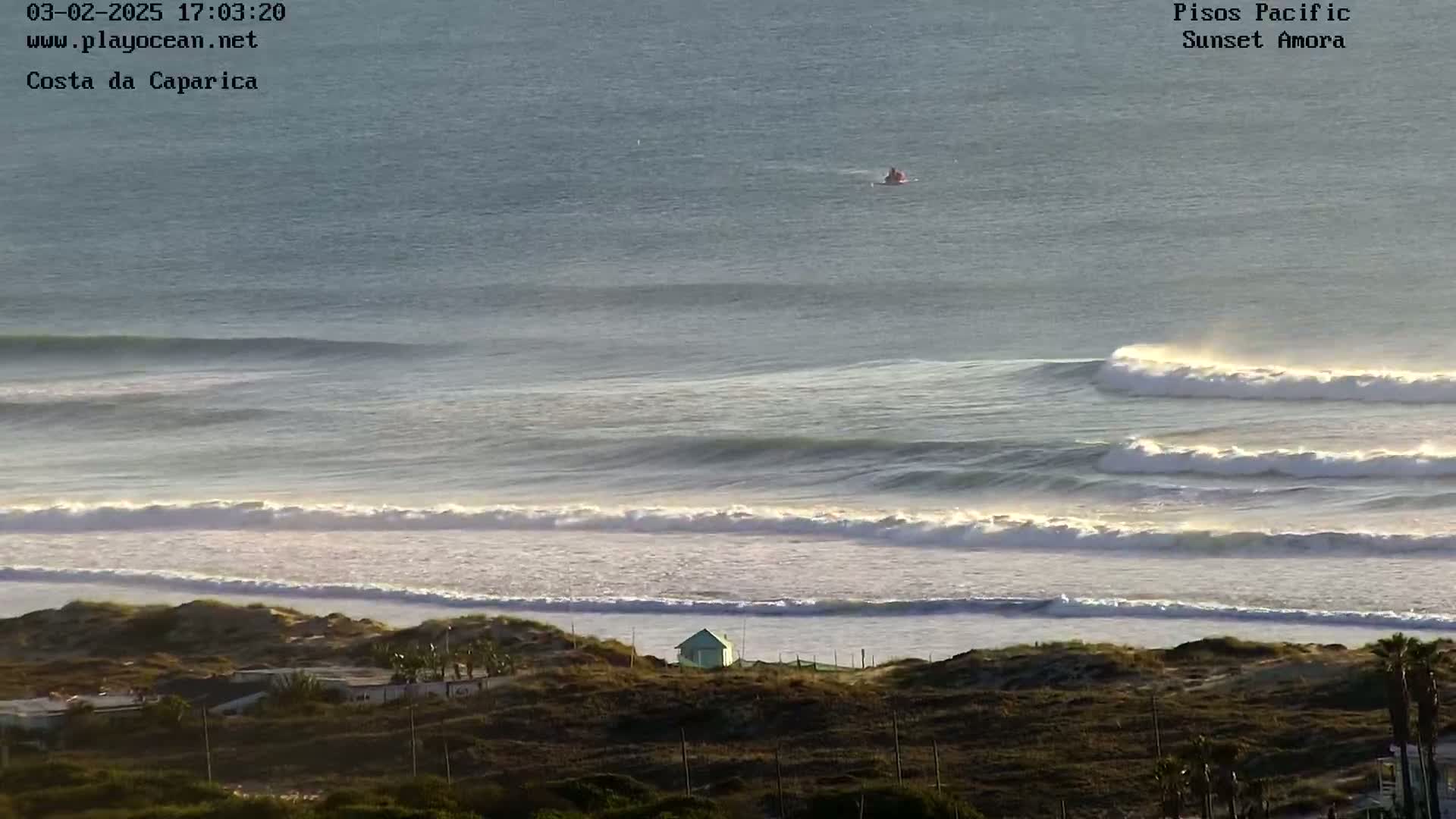A high-angle view shows a calm ocean with several long, rolling waves breaking on a sandy beach, overlooked by sandy dunes and a small building.