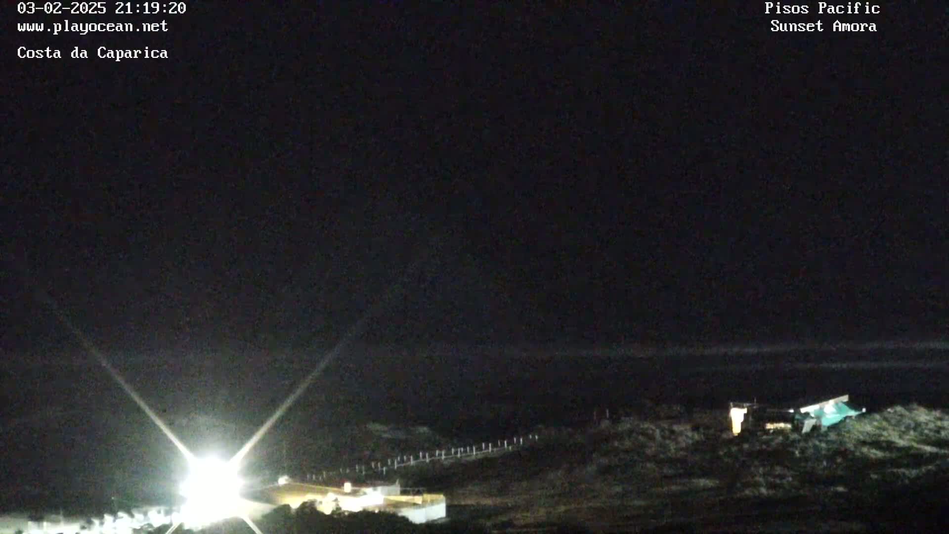 A nighttime coastal scene shows a dark ocean under a clear, black sky with bright lights illuminating a building and surrounding area.