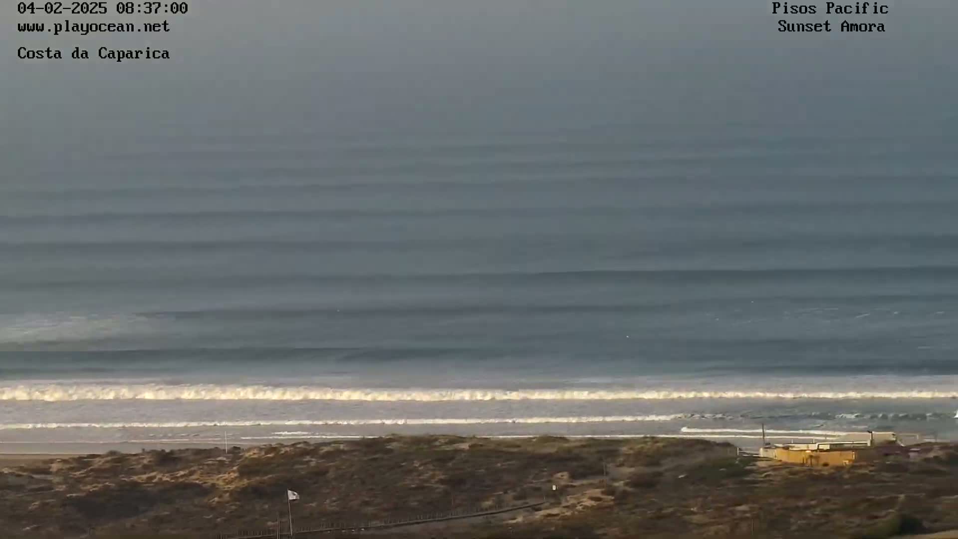 A long view of an ocean beach shows several lines of long, rolling waves approaching the shore on an overcast day.