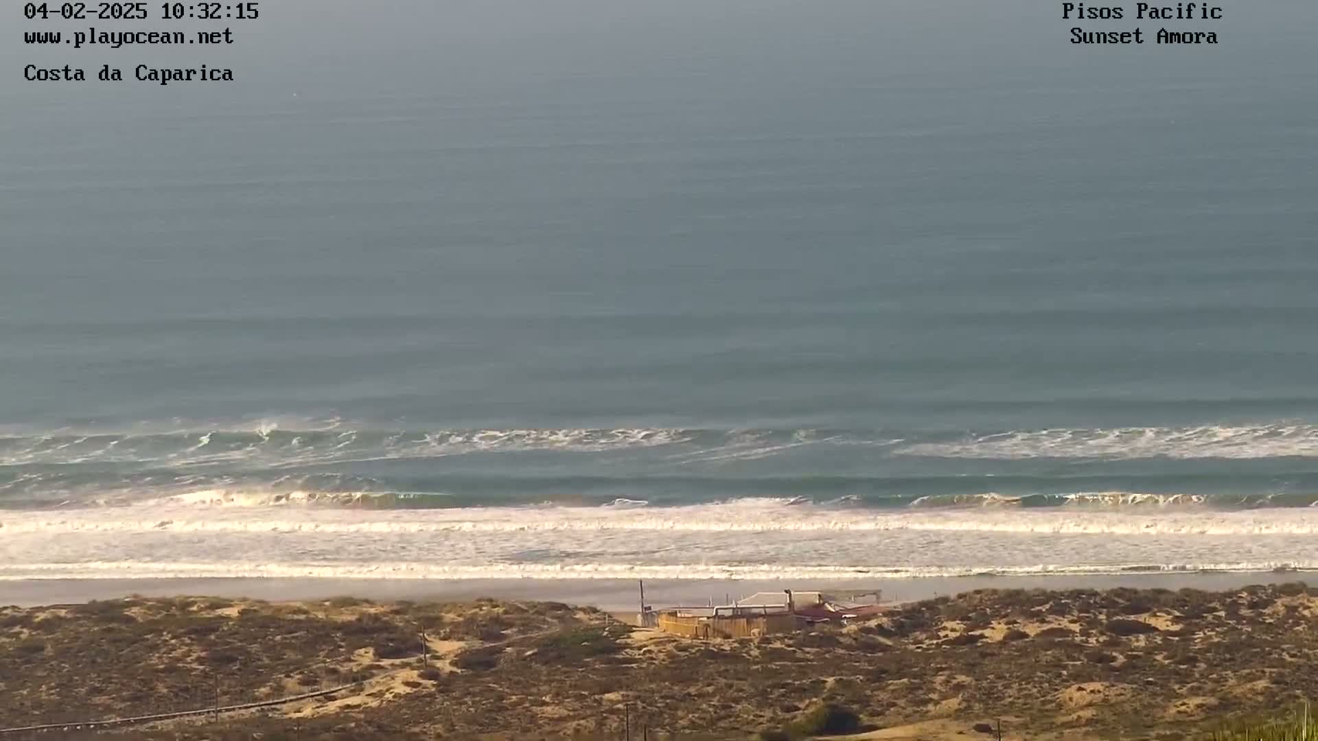 A long sandy beach with small waves breaking on the shore is seen from a high vantage point, under a mostly clear sky.