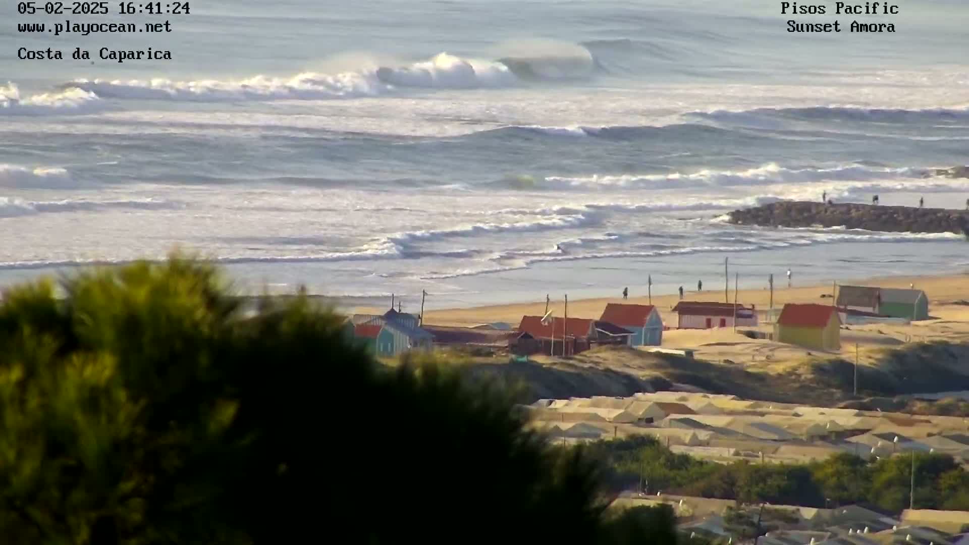 A view from a hilltop shows a sandy beach with colorful small buildings, a rocky jetty extending into a wavy ocean under a bright, mostly clear sky.