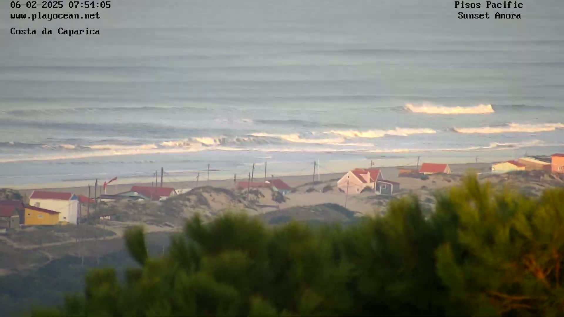 A coastal scene shows a sandy beach with small houses behind sand dunes, overlooked by green shrubbery, all under an overcast sky with waves breaking in the ocean.