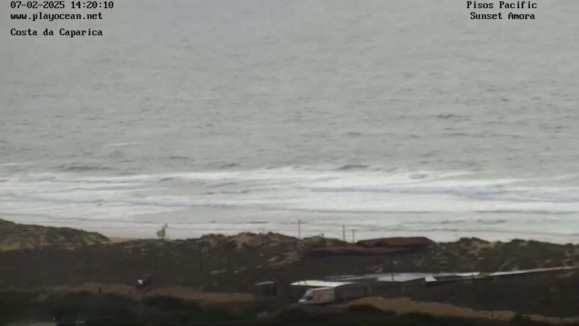 A long shot shows a beach with overcast skies and small waves rolling onto the shore, with some buildings and what appears to be a vehicle visible in the lower right.