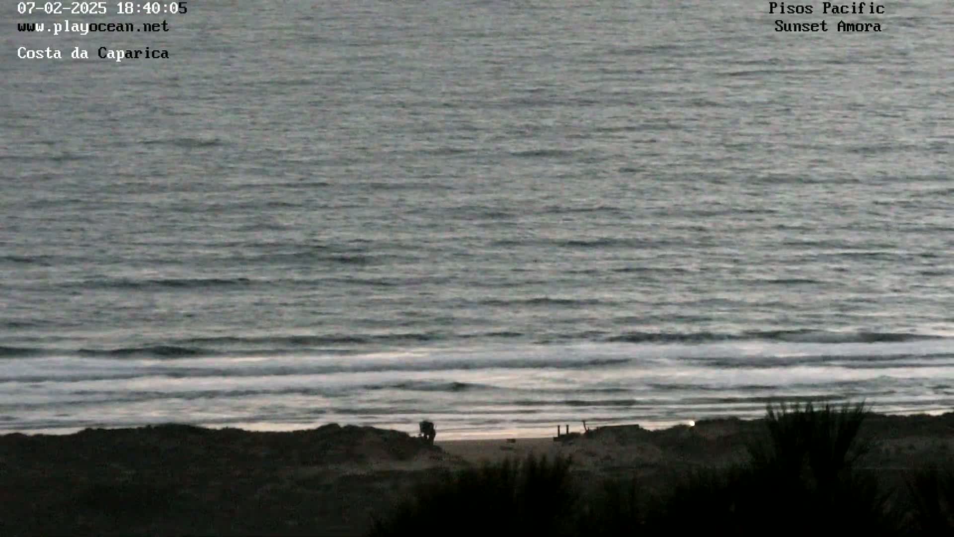 A dark, overcast view of a beach at dusk shows gentle waves lapping the shore.