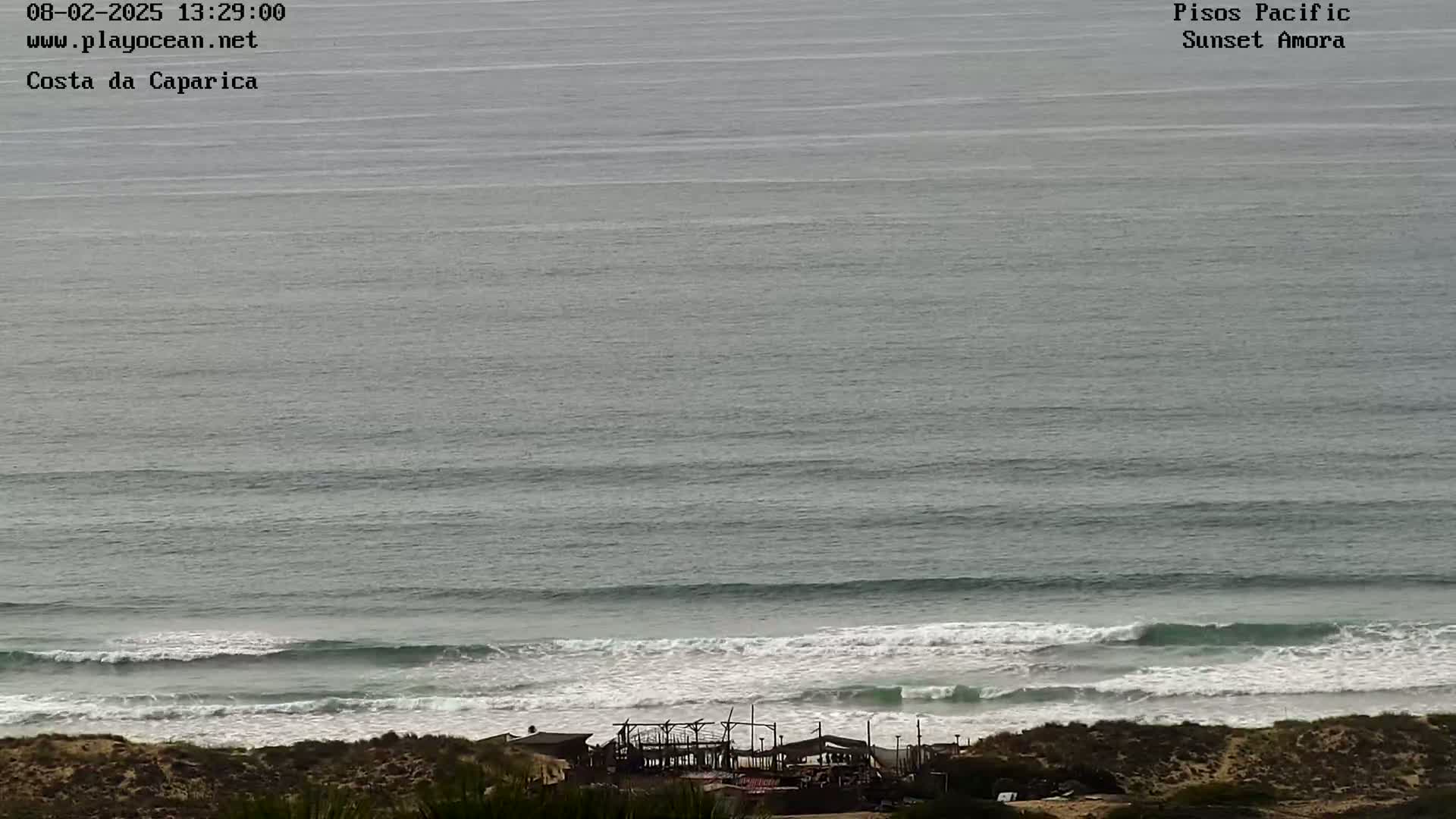 A high-angle, long shot shows a beach with a partially destroyed structure near the water's edge, under an overcast sky, with gently rolling waves in the ocean.