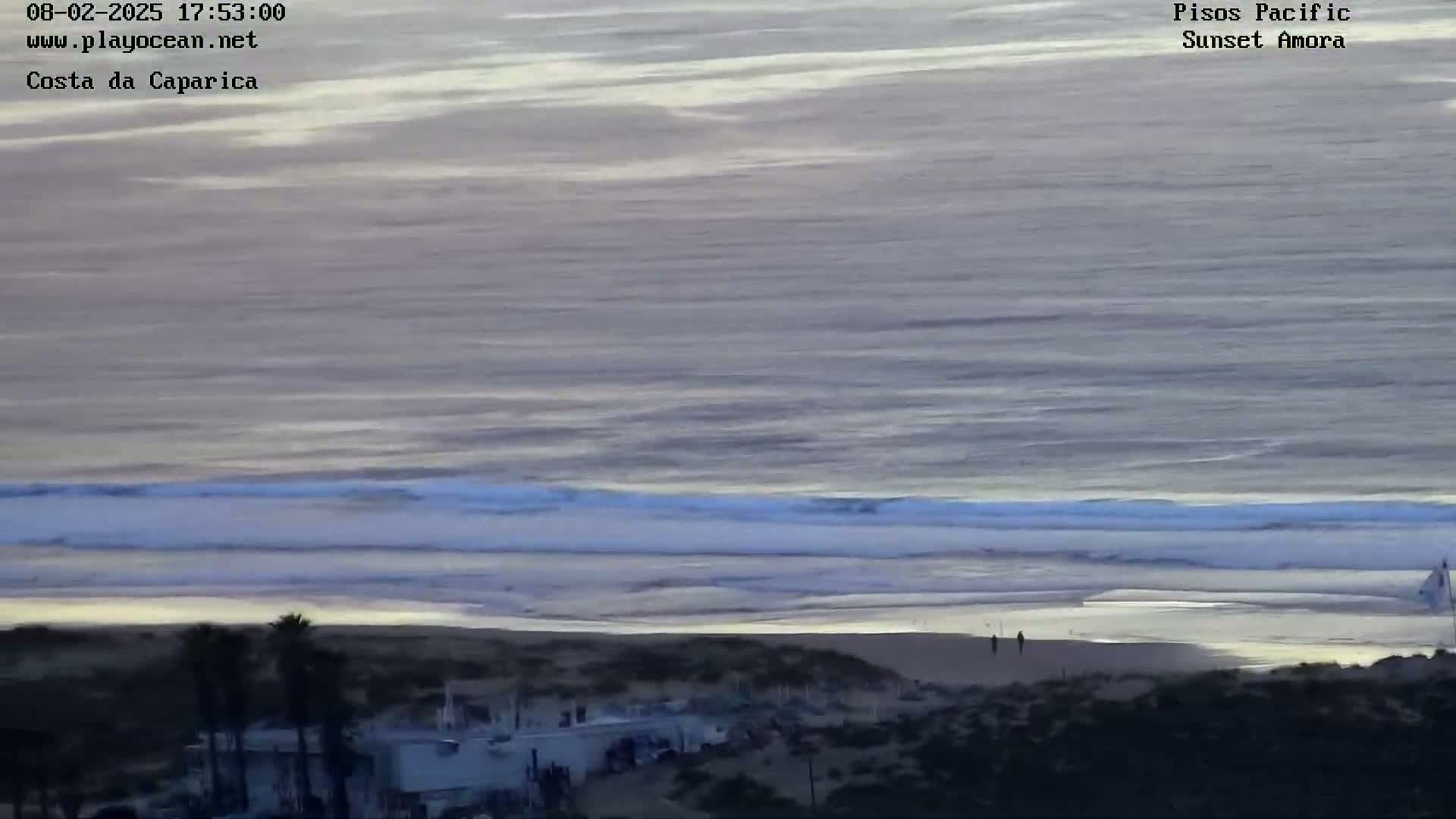 A beach scene at sunset shows calm ocean waves lapping a sandy shore, with a few people visible on the beach and buildings in the foreground.
