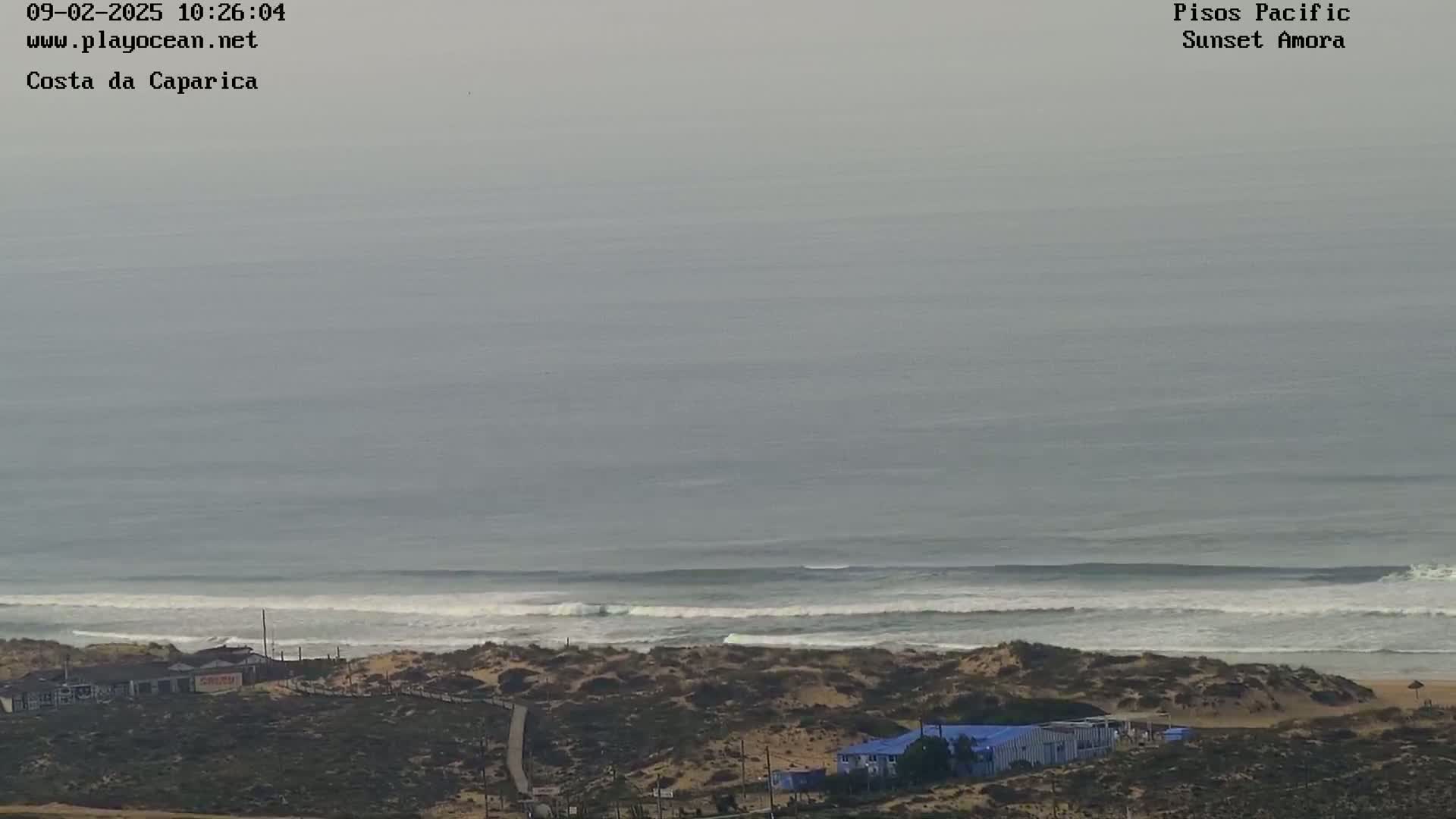 A hazy, overcast day shows a long stretch of sandy coastline with small waves rolling into shore, and several buildings visible near the beach.