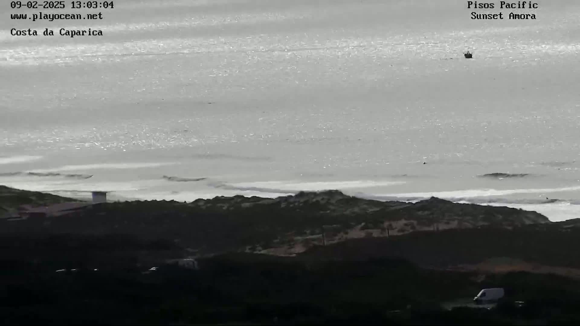 A mostly overcast day shows a distant boat on a calm ocean with small waves breaking on a rocky shoreline, and a van is visible in the lower right corner of the foreground.