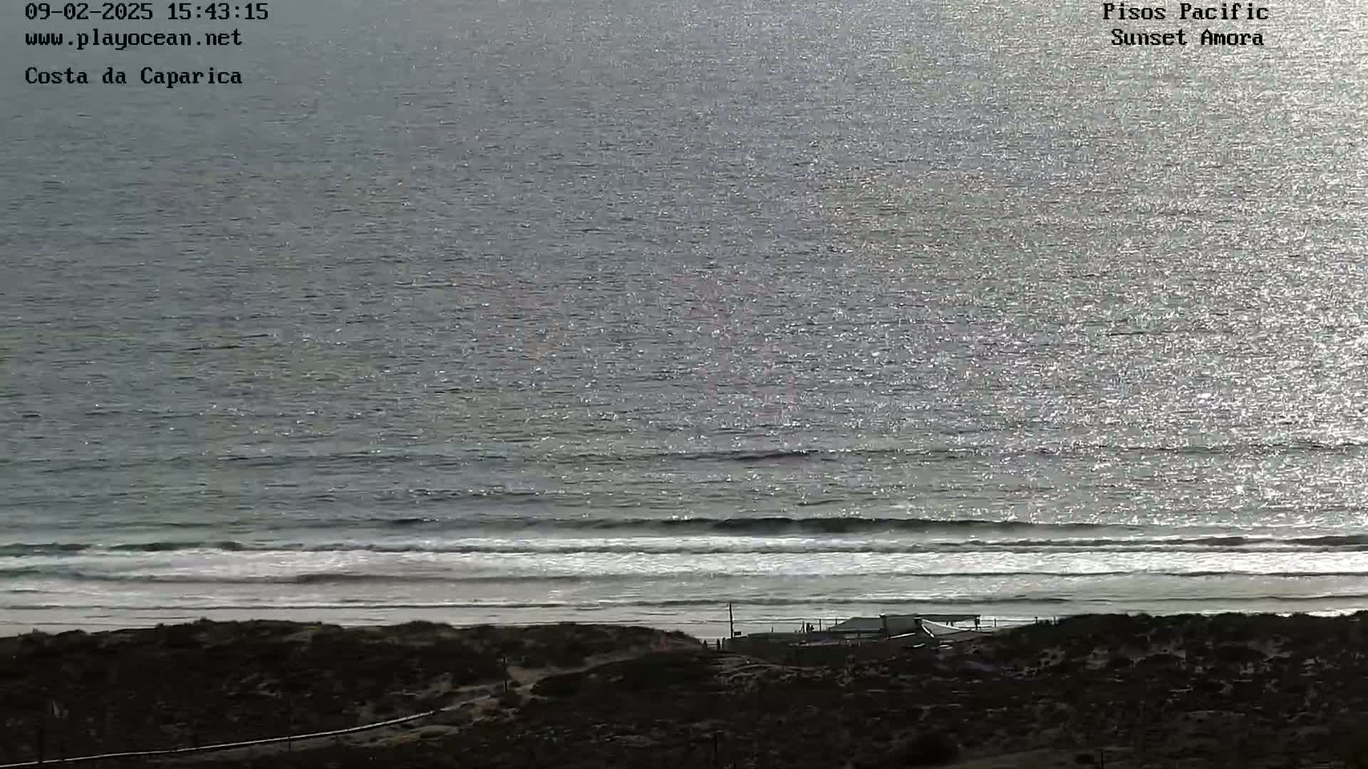 A sunny day shows a sparkling ocean with gentle waves rolling onto a sandy beach, where a few structures are visible near the shoreline.