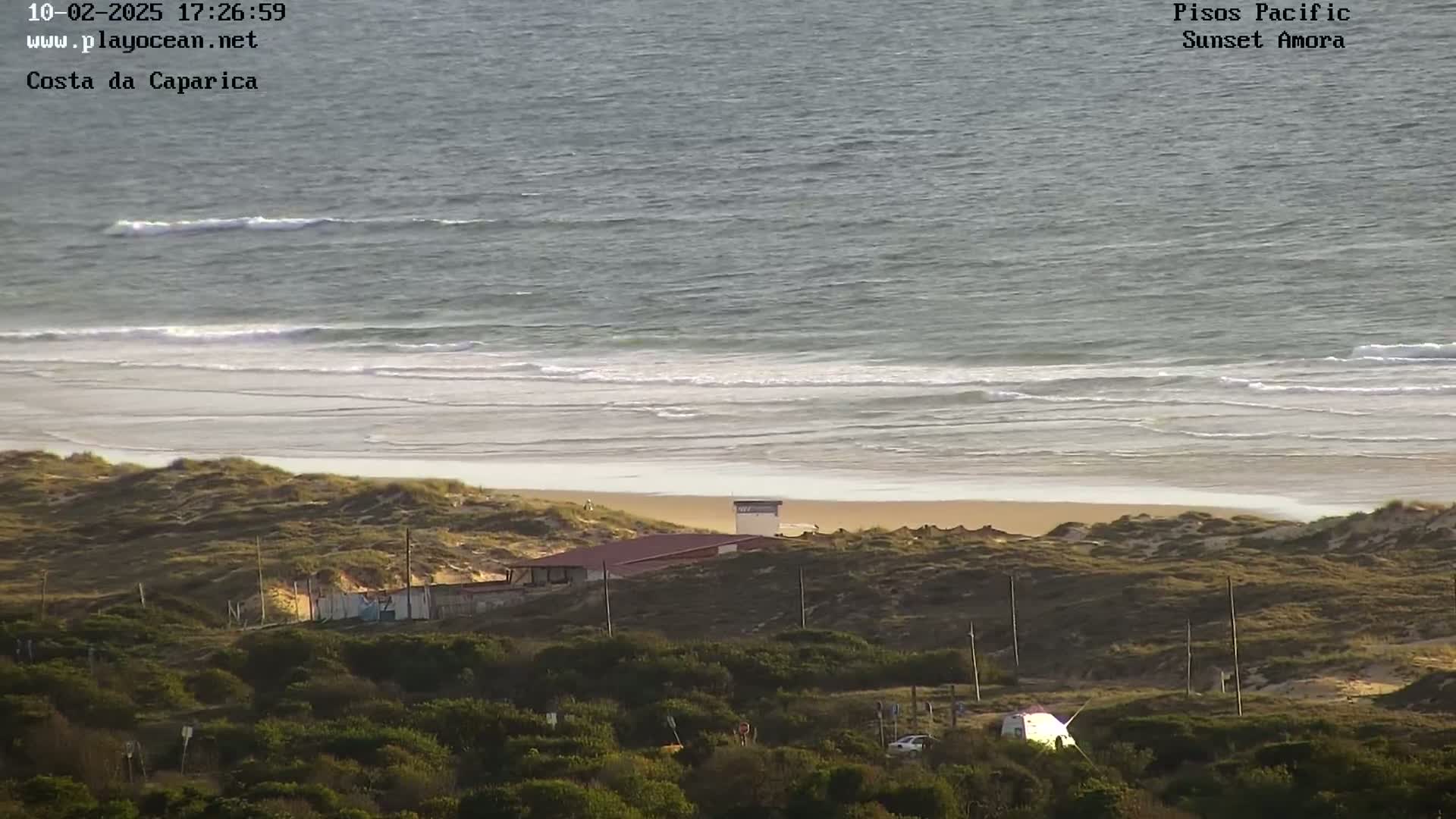 A sandy beach with gentle waves under a mostly clear sky is seen from a high vantage point overlooking low-lying vegetation and a small building.