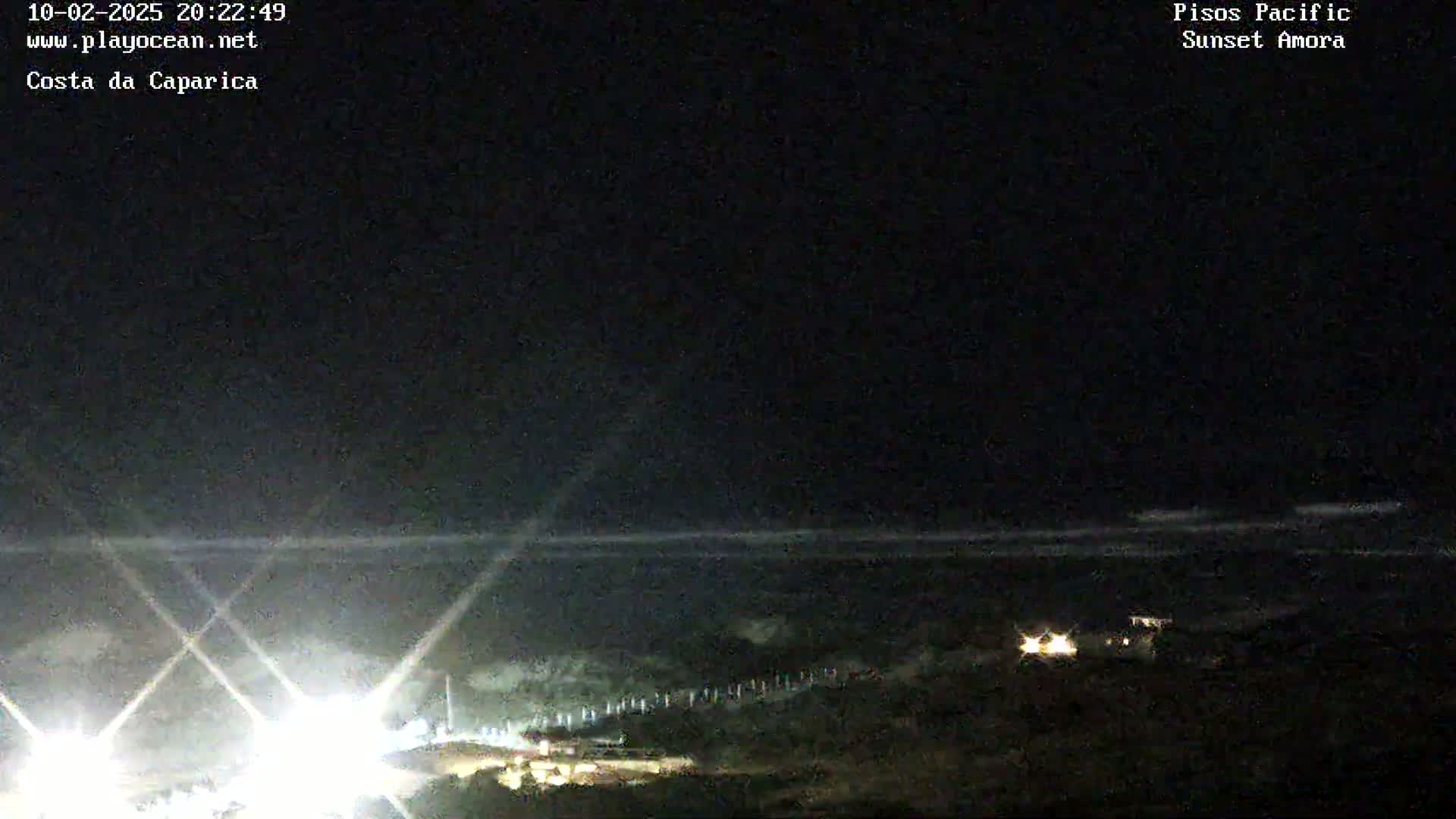 A nighttime coastal scene shows a dark ocean with a faint white line of waves, bright lights near the shore, and a dark, mostly featureless landmass.