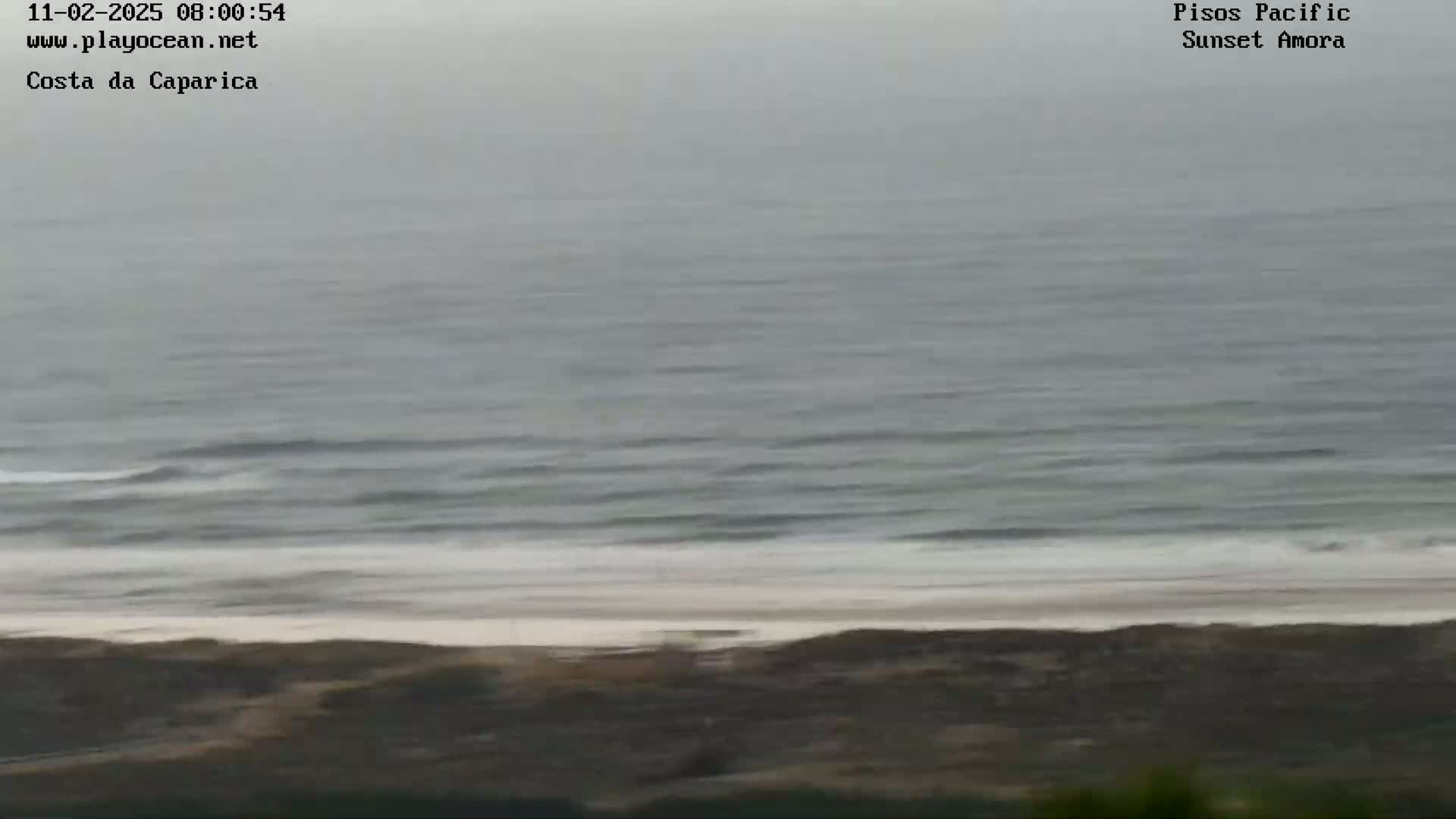 A mostly overcast, gray day shows a wide expanse of ocean with small waves breaking on a sandy beach.
