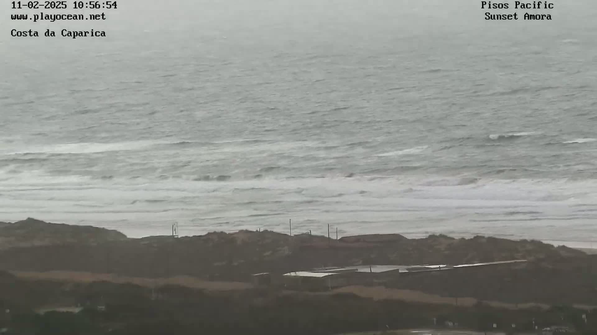 A hazy, overcast day shows a choppy ocean meeting a sandy shoreline with low-lying buildings visible in the foreground.