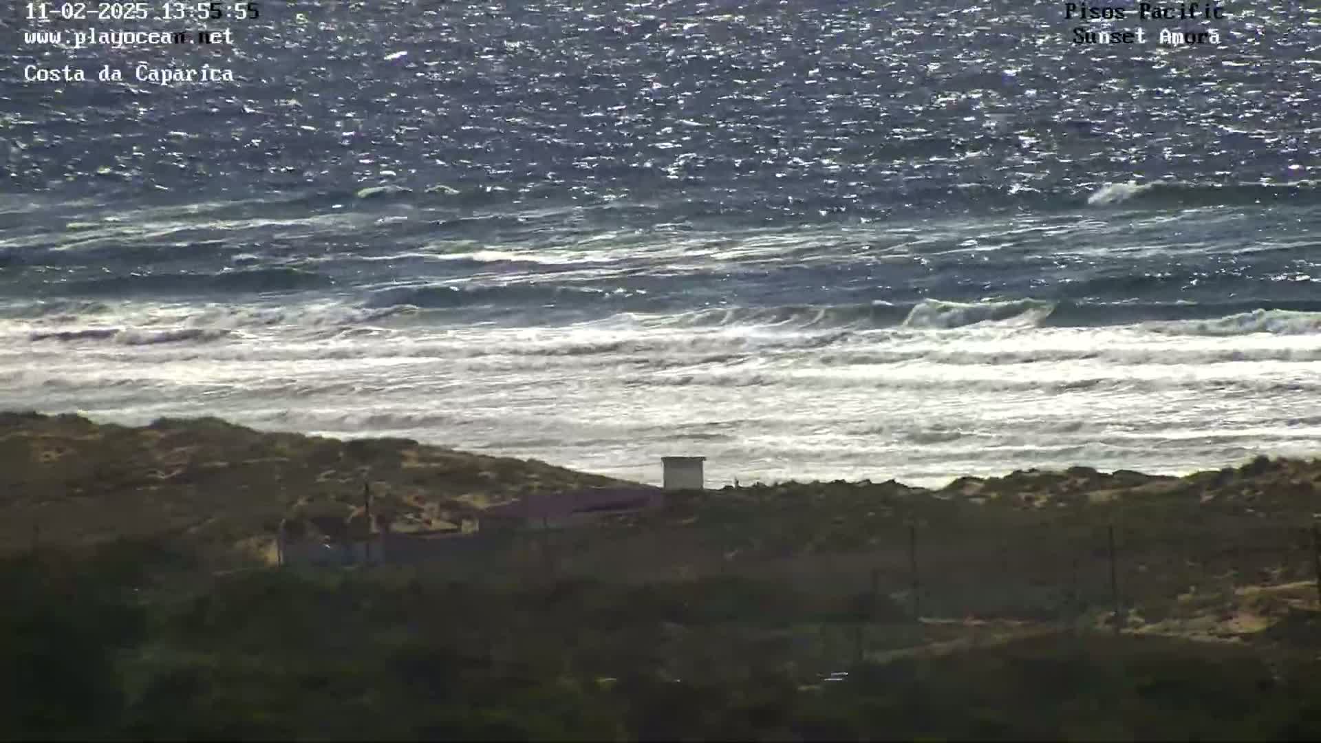 A somewhat overcast day shows a choppy ocean with waves breaking on a sandy shore, behind which sits low-lying vegetation and a few structures.