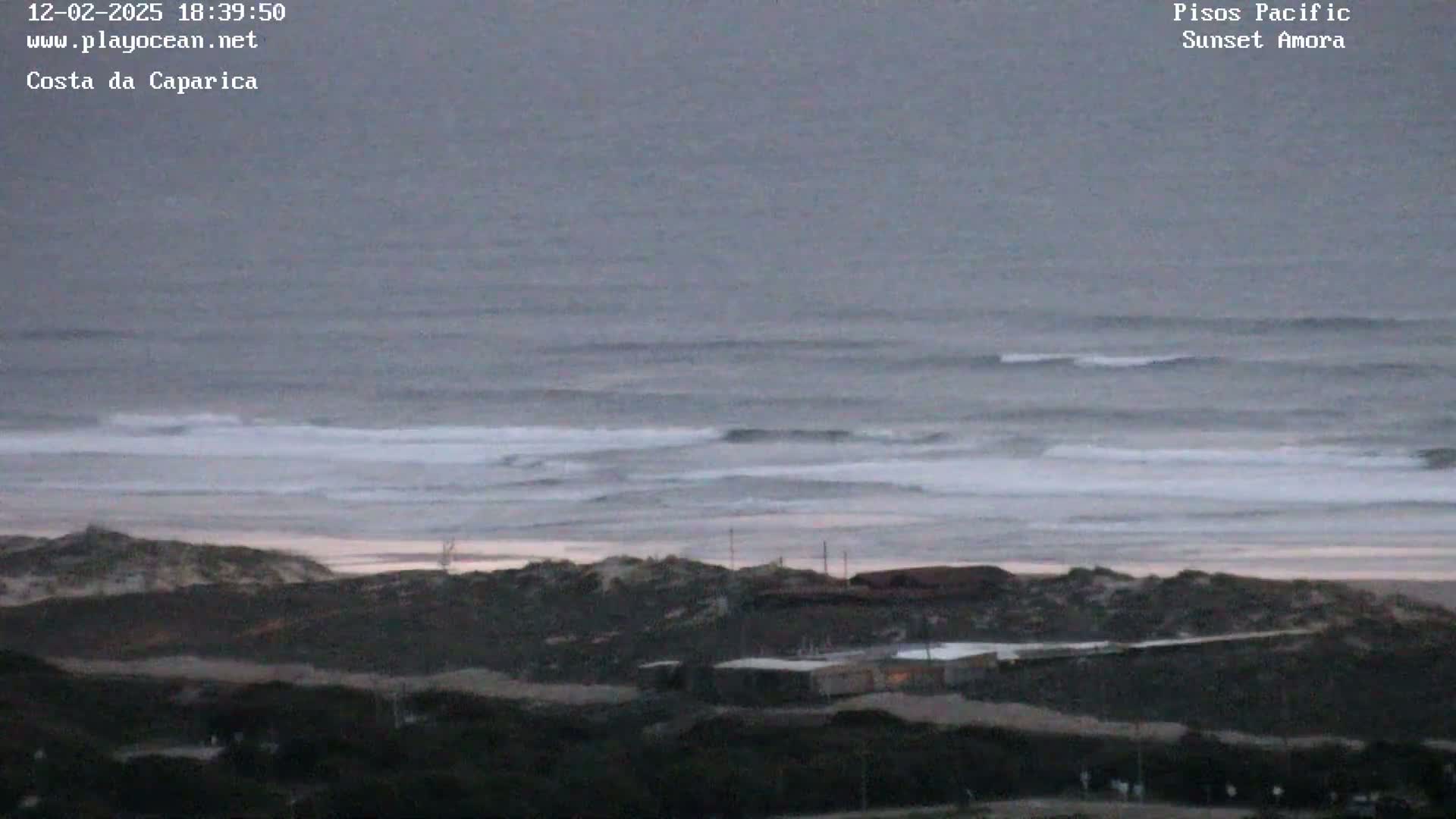 A gray, overcast sky overlooks a beach with gently rolling waves and dark, sandy dunes leading to some low-lying buildings.