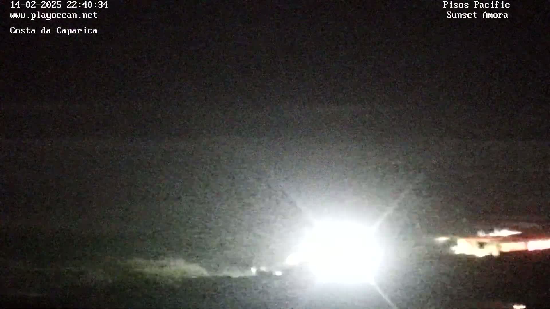 A bright light illuminates a dark, nighttime scene near a coastline, with some indistinct structures visible in the distance.
