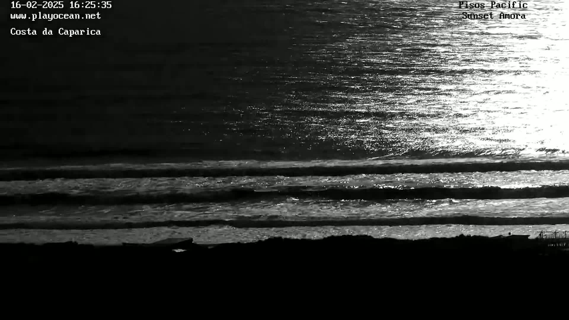 A grayscale image shows three parallel lines of small waves rolling onto a dark shoreline under a bright, possibly sunset sky.