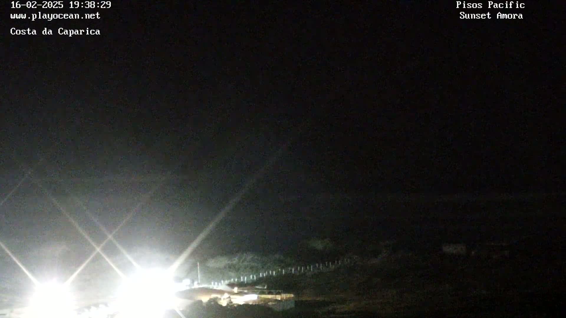 A nighttime, dark view shows bright lights from vehicles on a road near a fence, under a mostly clear sky.