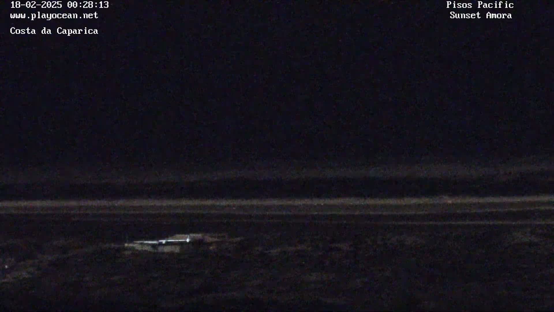 A dark, nighttime view shows a dimly lit shoreline with a faint horizontal line suggesting the ocean under a mostly clear sky.