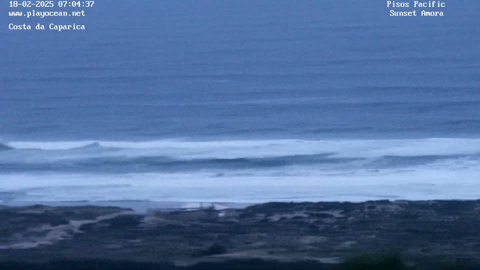 A high-angle, long shot shows a dark blue ocean with rolling waves breaking on a dark-colored shoreline under an overcast sky.