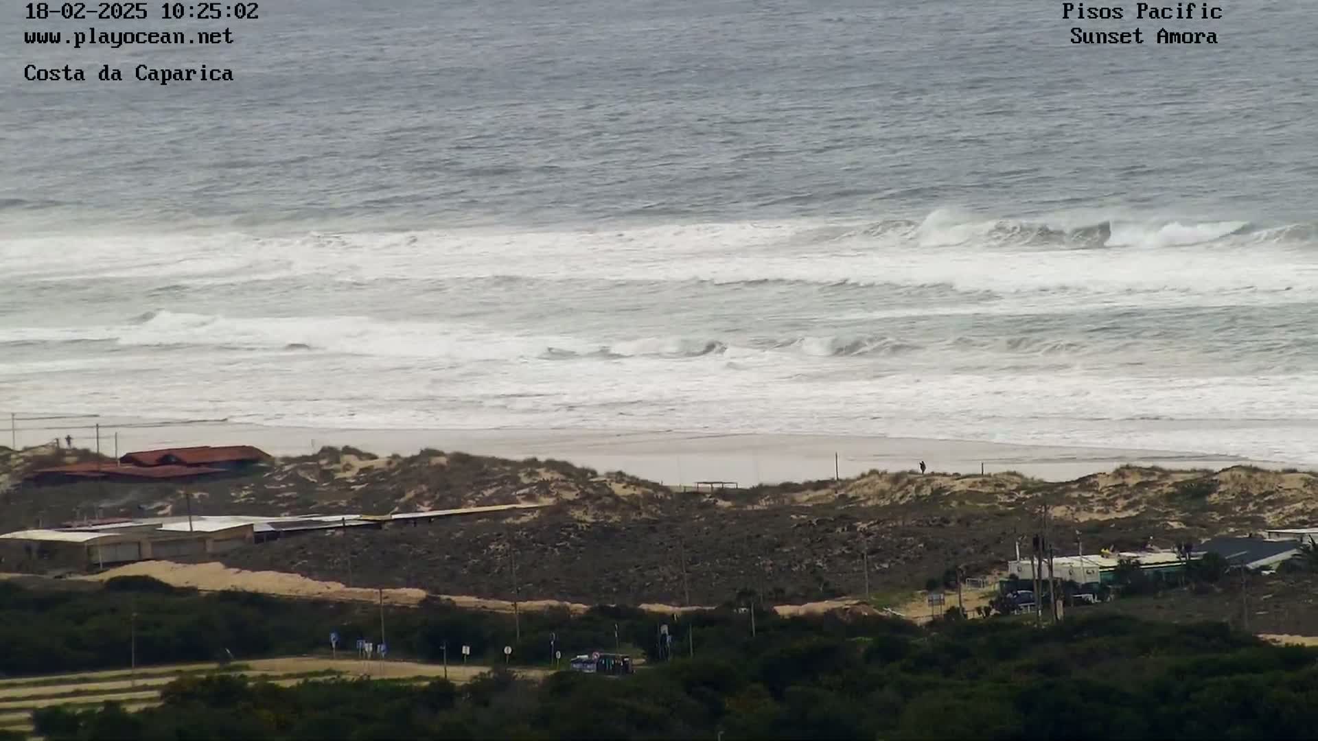 A beach with moderately rough waves and several buildings is seen from a distance, with dark-colored sand dunes separating the ocean from the buildings and vegetation.