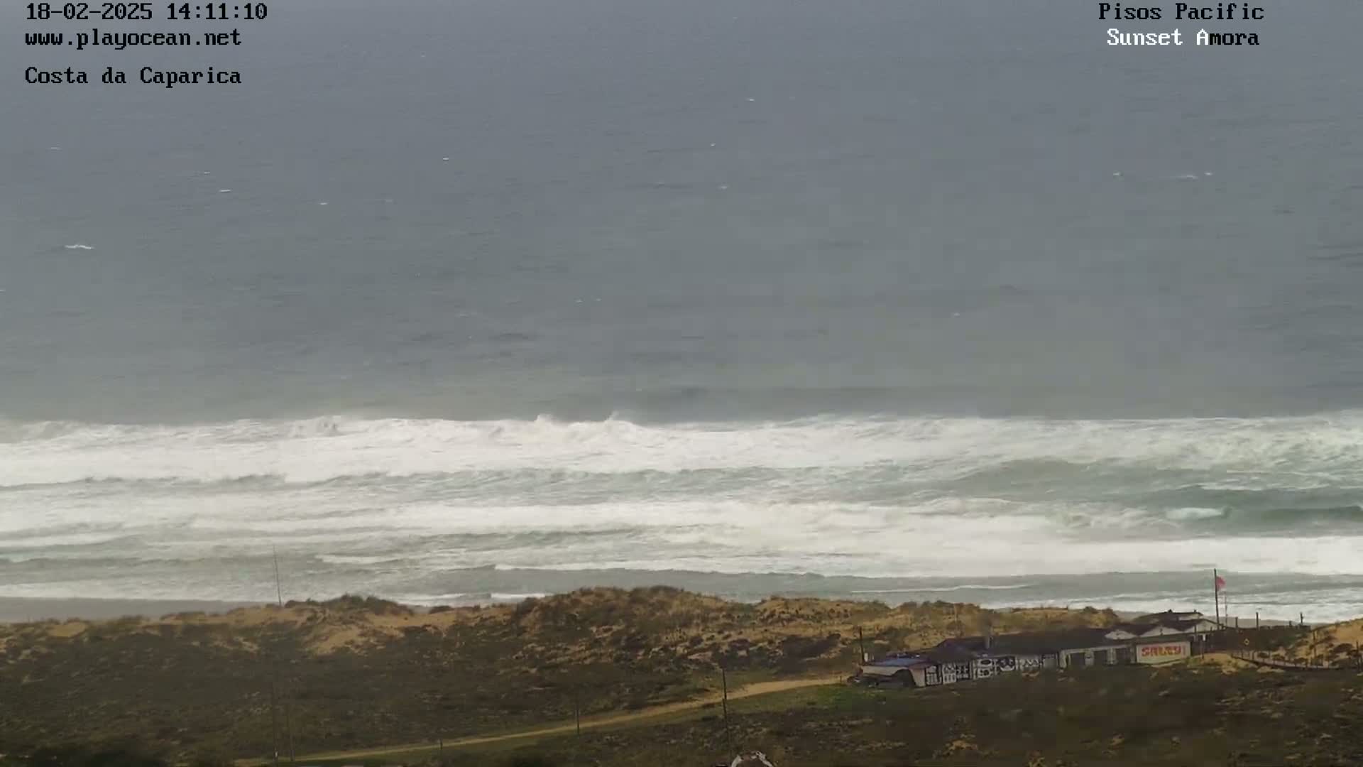 A high-angle view shows a beach with overcast skies and large waves crashing onto the shore, with a few buildings visible in the foreground.