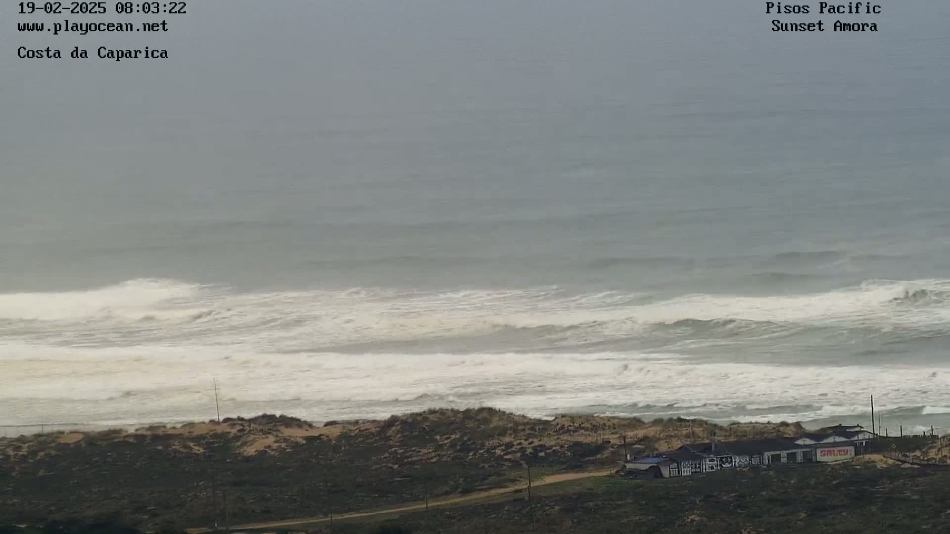 A high-angle, long shot shows a beach with large, white-capped waves crashing on the shore under an overcast sky.