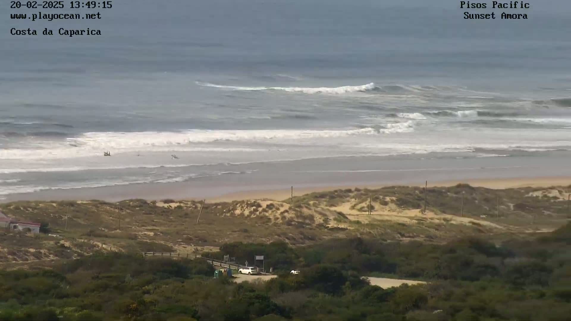 A beach with sand dunes and a few people in the ocean under an overcast sky, with waves breaking near the shore.