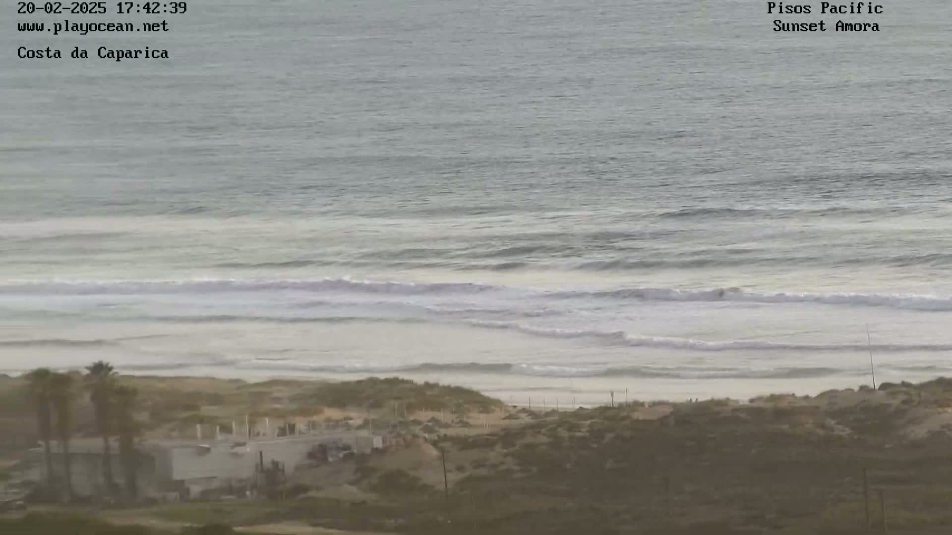 A hazy view shows a beach with moderate waves rolling onto the shore, under an overcast sky.