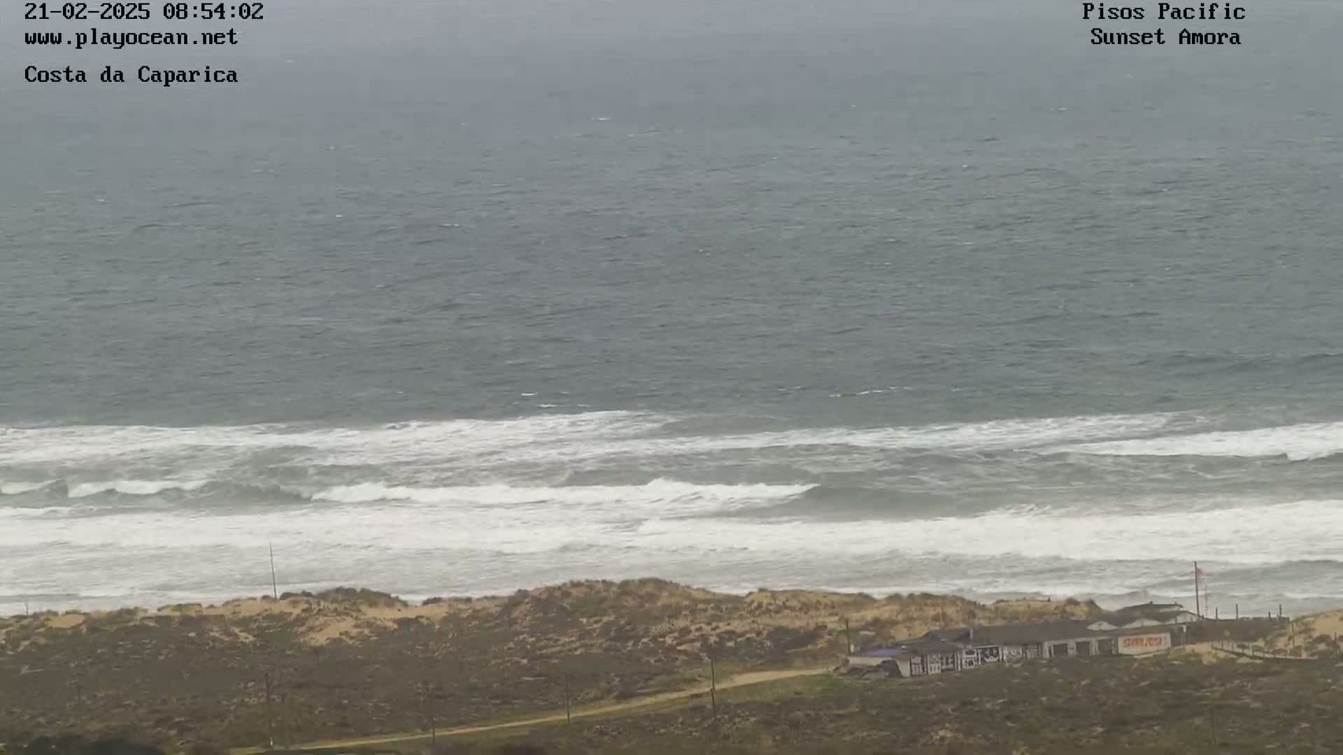 An overcast day shows a shoreline with small waves rolling into shore, and a few buildings are visible on the dune line.