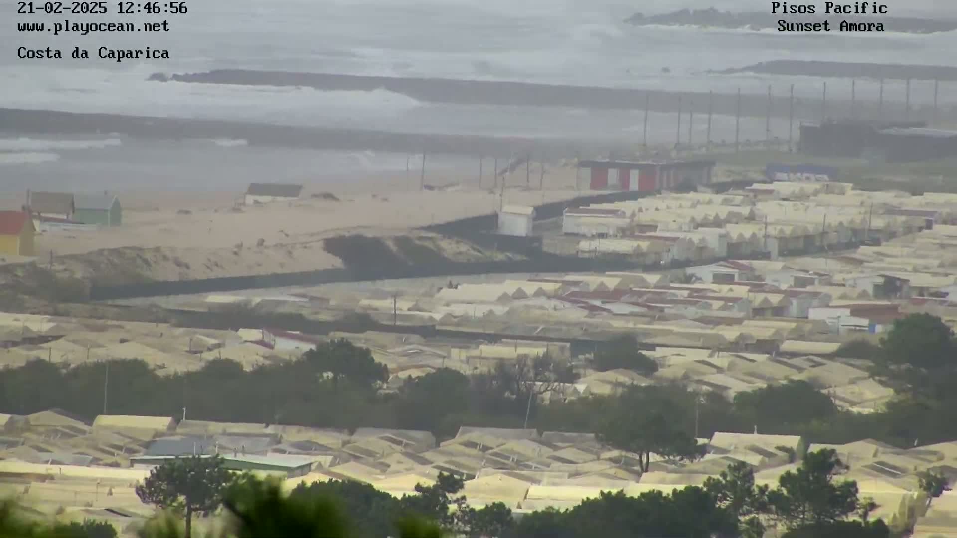 A hazy, overcast day reveals a coastal scene of a sandy beach with choppy waves, a line of buildings along the shore, and a large residential area stretching inland.
