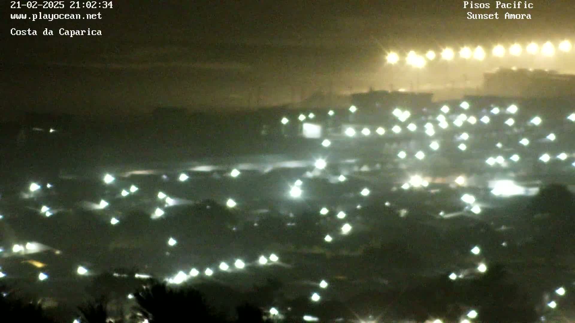 A hazy, nighttime view shows numerous scattered lights across a dark landscape, with brighter lights concentrated in the distance.