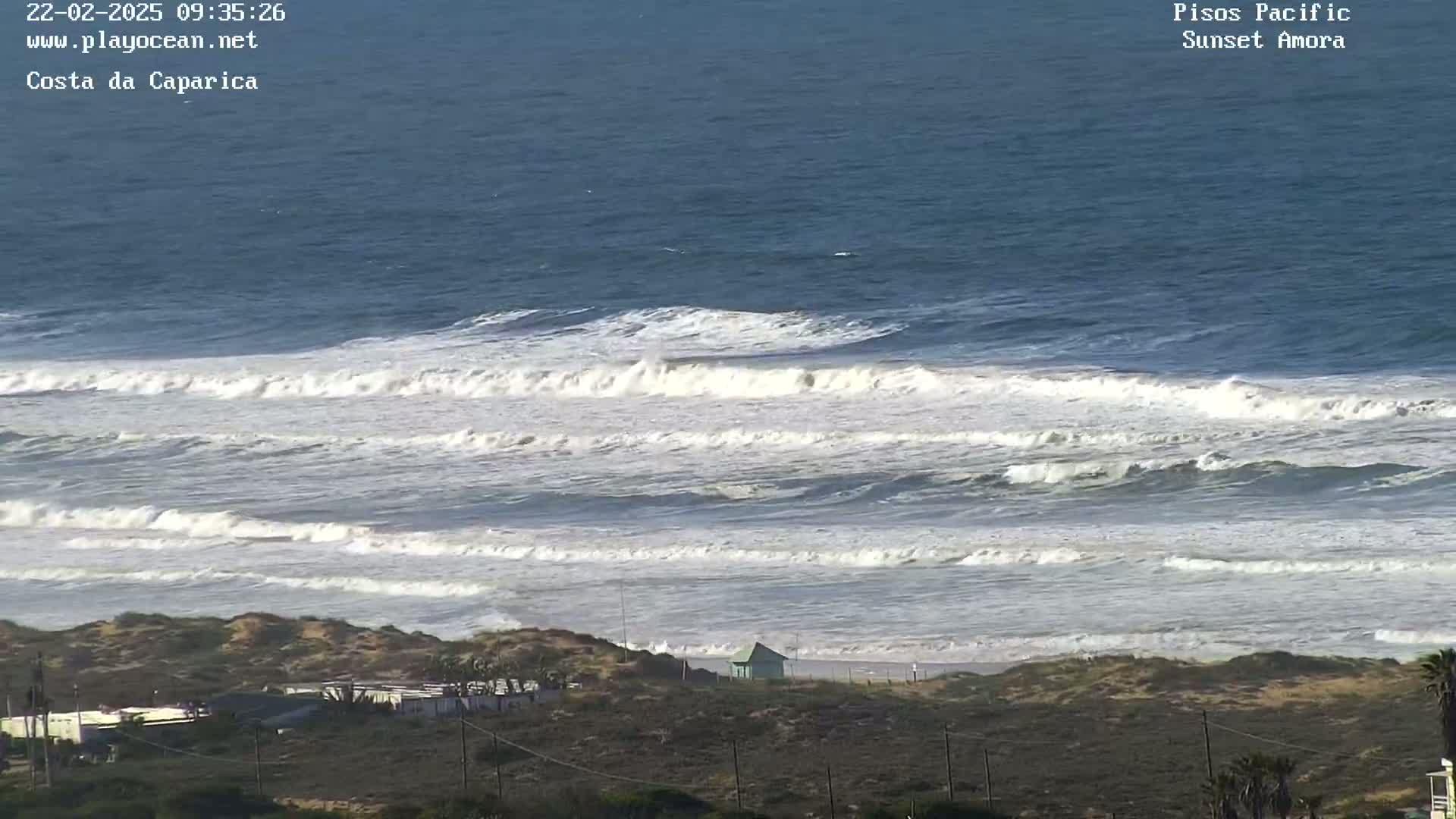 Ocean waves break on a sandy beach near a small building and sparse vegetation under a clear sky.