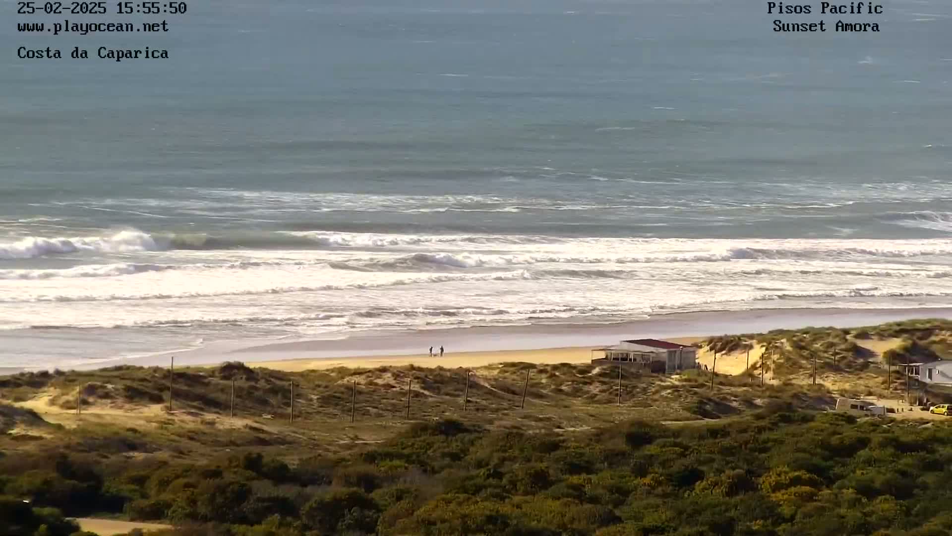 A sandy beach with some small buildings and people on it is backed by sand dunes and vegetation, overlooking a bright, wavy ocean under a mostly clear sky.