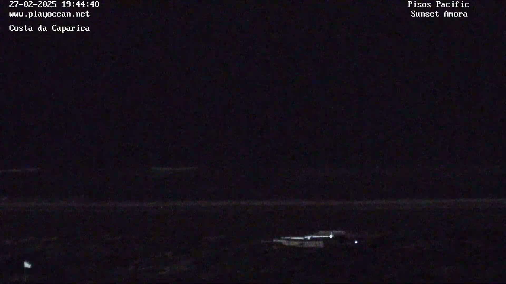 A dark nighttime coastal scene shows a mostly dark body of water with a faint horizon line and a few small lights visible near the bottom.