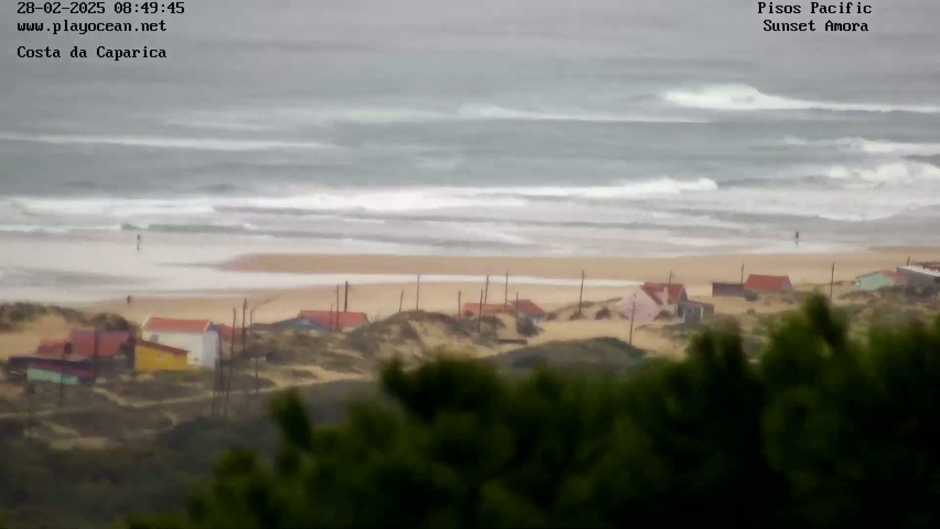 A view from above shows a beach with small houses nestled in sand dunes, and a wavy, gray ocean under an overcast sky.