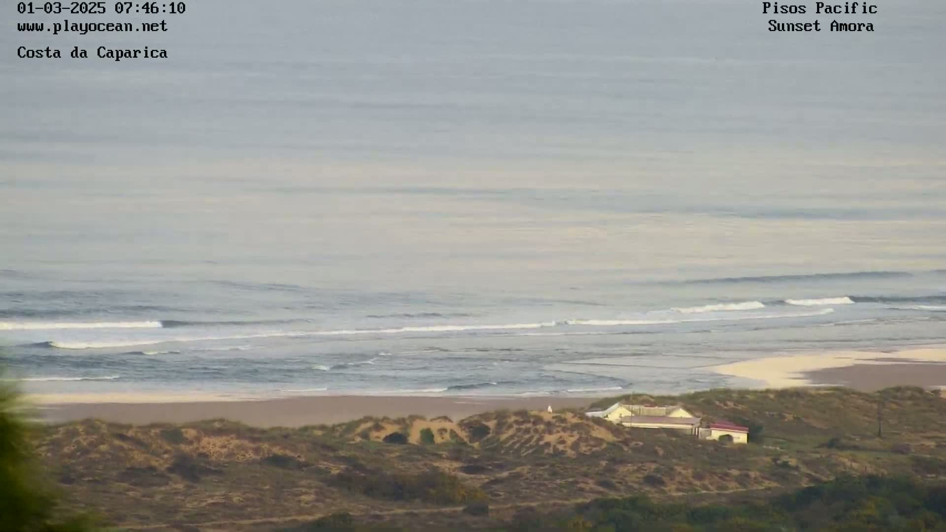 A long, sandy beach with a building on the dunes is visible beneath a hazy, overcast sky and a calm ocean with small waves breaking in the distance.