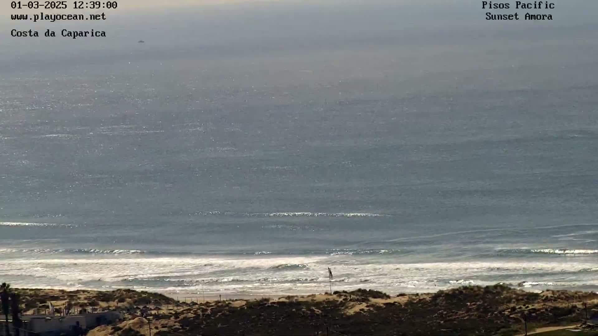 A mostly sunny, hazy day shows a wide expanse of ocean with waves breaking on a sandy shore.