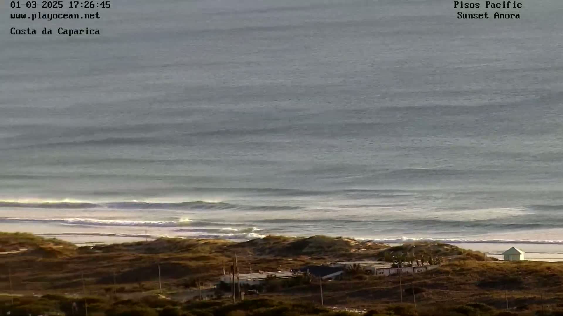 A long shot of an ocean beach with gentle waves under an overcast sky, showing a small building and some vegetation on the shore.