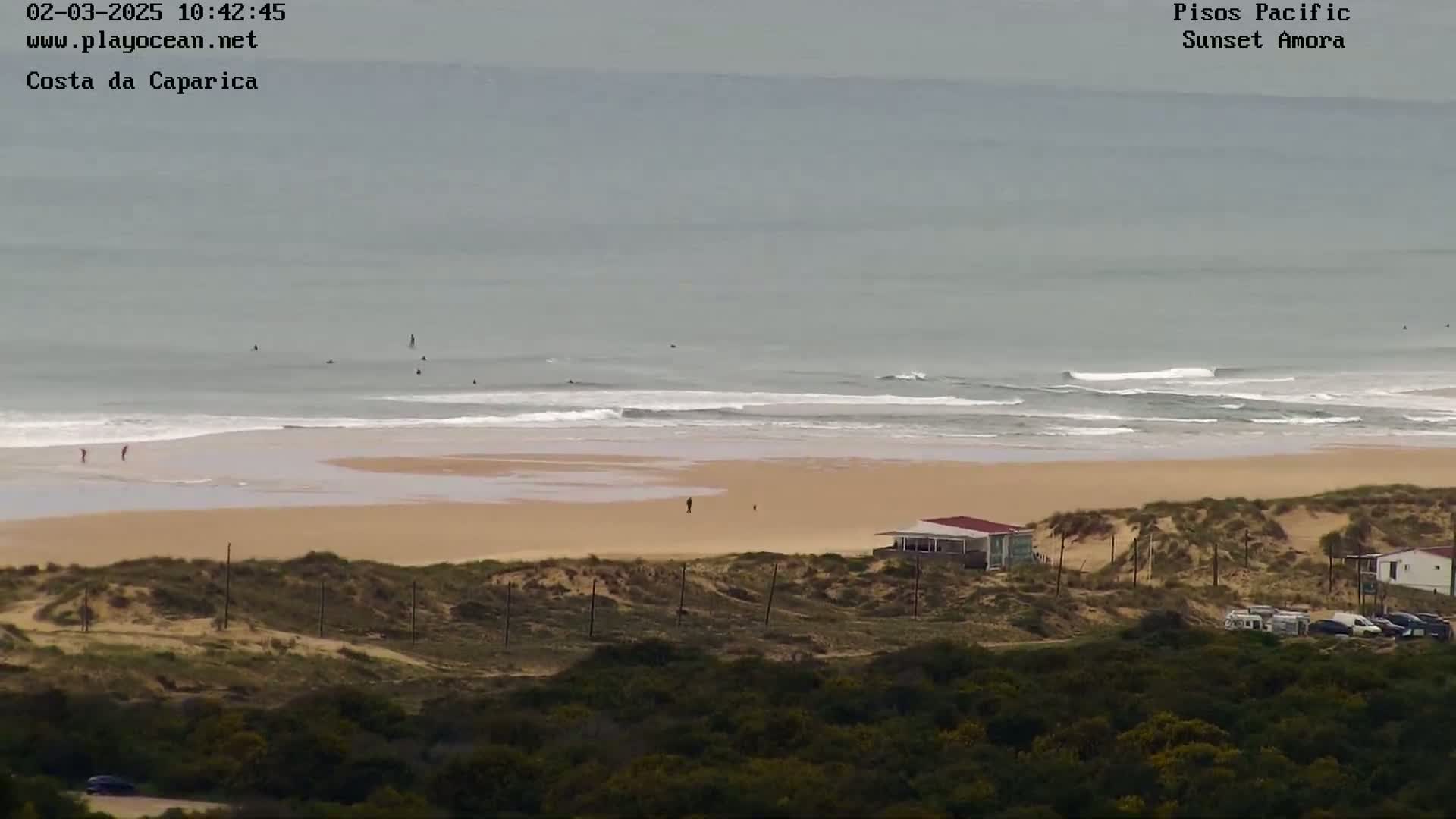An overcast day shows a beach with several surfers in the ocean, a sandy shoreline, and a small building near some dunes, all viewed from a high vantage point.