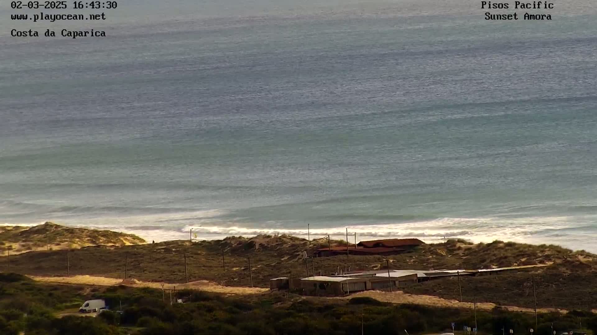 A coastal scene shows a calm ocean with small waves breaking on a sandy shore, behind which are some low-lying buildings and scrubland.